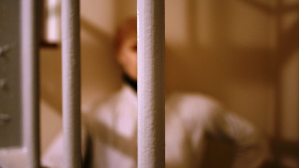 Oberkörper einer Frau in weißem Hemd verschwommen hinter Gitterstäben.